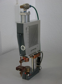 Eine kompakte Punktschweißzange mit pneumatischem Zylinder steht auf einem Tisch vor einer weißen Wand. Kupferne Elektroden und ein Anschluss für Druckluft sind sichtbar, das Gerät trägt eine Skalierung zur Kraft- oder Zeitmessung.