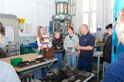 Fünf Jugendliche stehen an einem Arbeitstisch in einer Werkhalle, während ein Mann in Schutzkleidung eine Maschine erklärt. Auf dem Tisch liegen mehrere grüne Bauteile und Werkzeuge, im Hintergrund sind weitere Maschinen und technische Anlagen sichtbar.