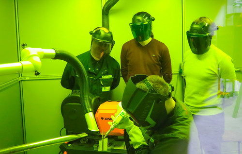 ine Gruppe von Personen mit Schweißhelmen beobachtet eine Laserschweißarbeit, die in einer mit grünem Schutzglas ausgestatteten Kabine durchgeführt wird.