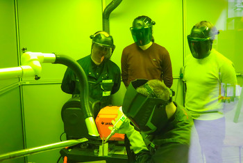 ine Gruppe von Personen mit Schweißhelmen beobachtet eine Laserschweißarbeit, die in einer mit grünem Schutzglas ausgestatteten Kabine durchgeführt wird.