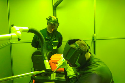 ine Gruppe von Personen mit Schweißhelmen beobachtet eine Laserschweißarbeit, die in einer mit grünem Schutzglas ausgestatteten Kabine durchgeführt wird.