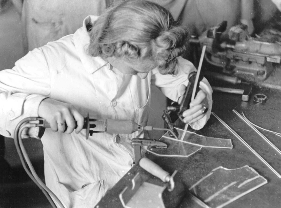 Schwarz-weiß-Fotografie einer Frau in Arbeitskittel beim Schweißen von Kunststoffteilen an einem Werktisch, konzentriert führt sie eine Flamme und ein Werkzeug an ein eingespanntes Werkstück.