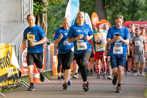 Vier Läufer (drei Männer, eine Frau) in blauen Laufshirts überqueren die Start-Ziel-Linie eines Firmenlaufs, hinter ihnen stehen weitere Läufer bereit. Der Bereich ist mit Werbebannern und Bäumen gesäumt.