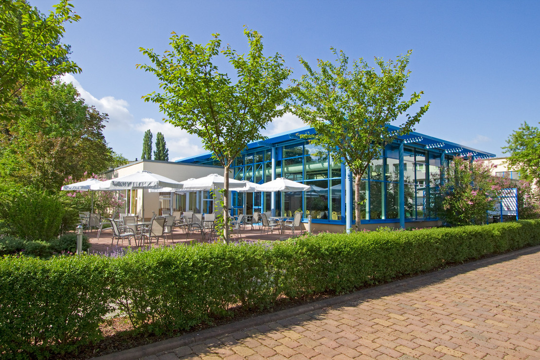 Außenansicht eines verglasten Veranstaltungsbereichs mit blauen Stahlträgern. Davor eine Terrasse mit mehreren Sitzgruppen und weißen Sonnenschirmen, umgeben von blühenden Sträuchern und jungen Bäumen. Im Vordergrund eine gepflegte Hecke und gepflasterter Weg.