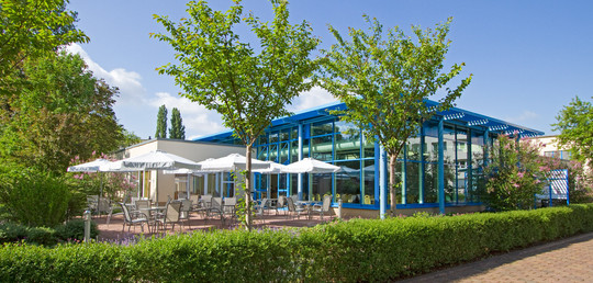 Außenansicht eines verglasten Veranstaltungsbereichs mit blauen Stahlträgern. Davor eine Terrasse mit mehreren Sitzgruppen und weißen Sonnenschirmen, umgeben von blühenden Sträuchern und jungen Bäumen. Im Vordergrund eine gepflegte Hecke und gepflasterter Weg.