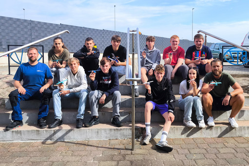 Eine Gruppe von Jugendlichen flankiert von zwei Männern sitzt auf einer niedrigen Betontreppe im Freien und blickt in die Kamera. Alle halten ein Flugzeugmodell aus dem 3D-Drucker in der Hand. Im Hintergrund sind Gebäude, Geländer und technische Anlagen zu sehen.