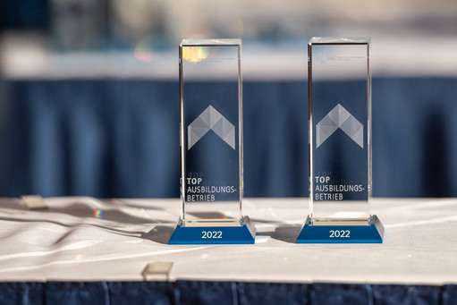 Zwei gläserne Trophäen mit der Aufschrift „Top Ausbildungsbetrieb 2022“ stehen auf einem weiß gedeckten Tisch vor blauem Hintergrund.