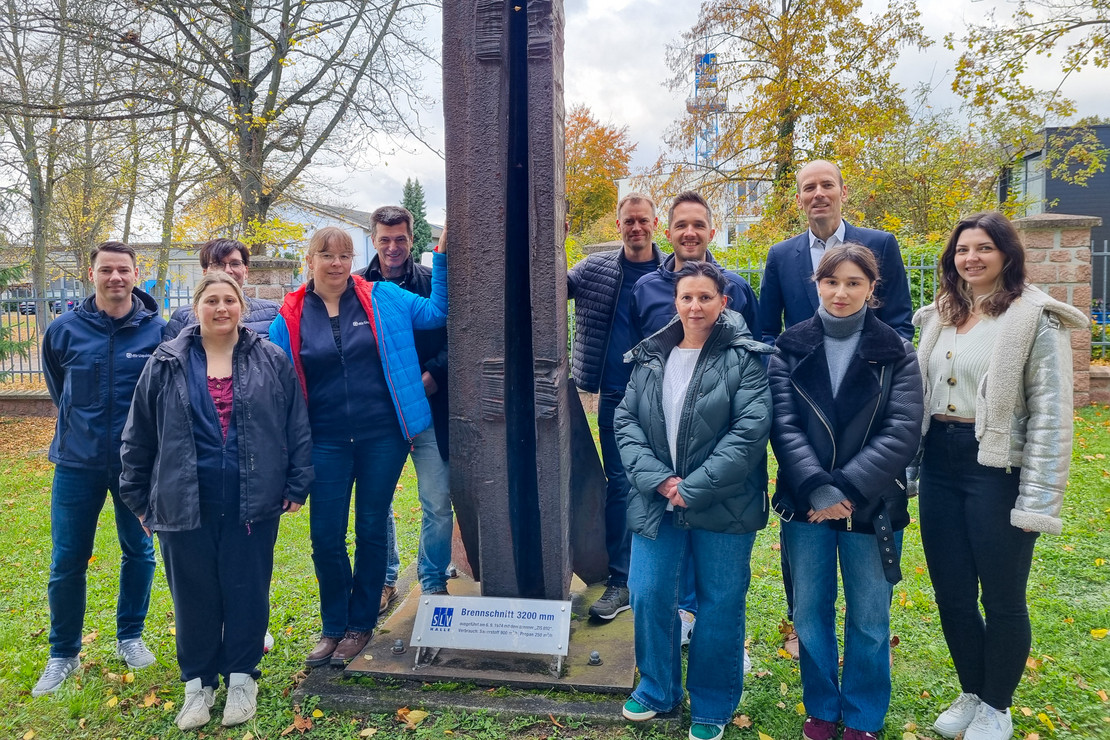 Neun Personen stehen im Freien vor einer hohen Metallskulptur und lächeln in die Kamera, im Hintergrund herbstliche Bäume und Gebäude.