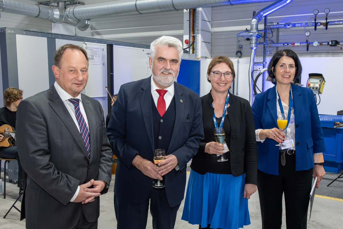 Zwei Männer und zwei Frauen in Businesskleidung stehen in einer Werkhalle mit Getränken in der Hand und lächeln in die Kamera; im Hintergrund sind Schweißtechnik und Lüftungsrohre sichtbar.