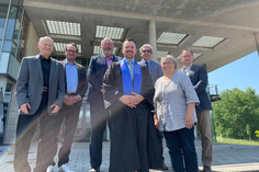 Sechs Männer und eine Frau posieren für ein Gruppenfoto. Mann in der Mitte trägt die Kleidung eines Promovierenden.