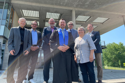 Sechs Männer und eine Frau posieren für ein Gruppenfoto. Mann in der Mitte trägt die Kleidung eines Promovierenden.