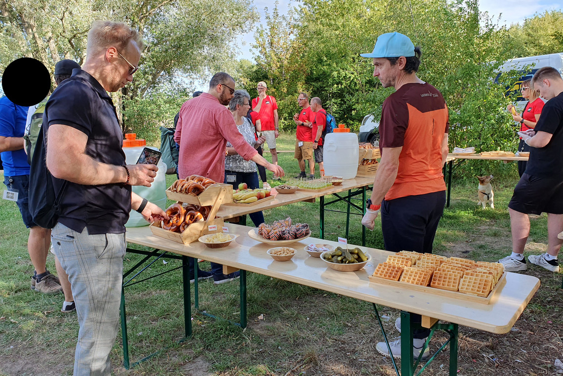 Teilnehmende eines Outdoor-Events bedienen sich an einem Buffet mit Laugengebäck, Waffeln, Gewürzgurken und Getränken auf Biertischen, im Hintergrund stehen weitere Personen in sportlicher Kleidung in einer Gartenlandschaft.