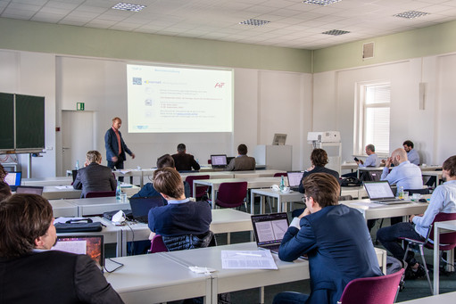Teilnehmende eines Fachseminars sitzen mit Laptops in einem hellen Seminarraum, ein Mann präsentiert stehend vor einer Leinwand mit projizierten Inhalten.