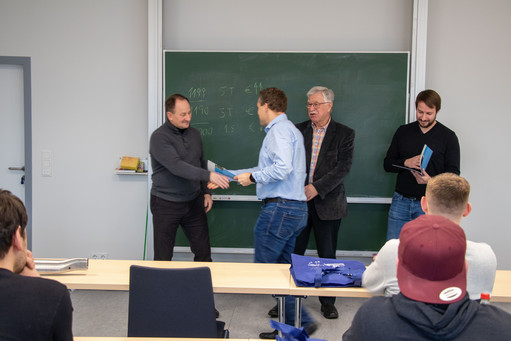 Vier Männer stehen vor einer Tafel in einem Seminarraum, einer überreicht einem anderen ein Dokument per Handschlag, im Vordergrund sitzen Zuhörer an Tischen