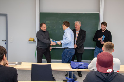 Vier Männer stehen vor einer Tafel in einem Seminarraum, einer überreicht einem anderen ein Dokument per Handschlag, im Vordergrund sitzen Zuhörer an Tischen