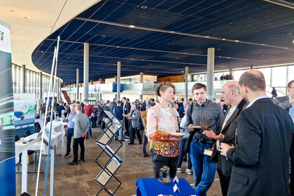 Besucherinnen und Besucher stehen in einer Messehalle vor einem Informationsstand der SLV Halle GmbH. Mehrere Personen führen Gespräche am Stand mit blauem DVS SLV Halle Tischtuch. Im Hintergrund sind weitere Messestände, Roll-ups und eine große Decke mit Beleuchtung zu sehen.