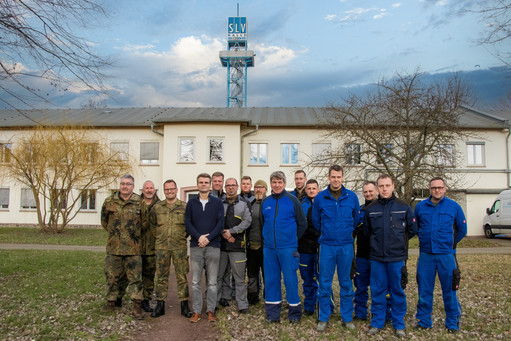 Gruppenfoto von vierzehn Männern in Arbeitskleidung oder Bundeswehr-Uniform vor einem weißen Gebäude mit dem blauen SLV-Prüfturm im Hintergrund unter bewölktem Himmel