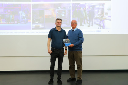 Zwei Männer stehen vor einer projizierten Präsentationsfolie auf einer Bühne und halten ein Buch. Sie befinden sich in einem Vortragsraum mit Leinwand im Hintergrund, die das DVS-Schaudepot zeigen.