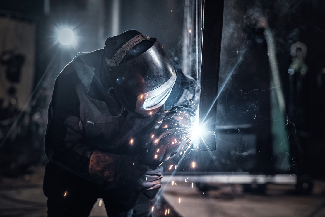 Schweißer mit Schutzhelm arbeitet an einem Werkstück, Funken sprühen hell in dunkler Umgebung.