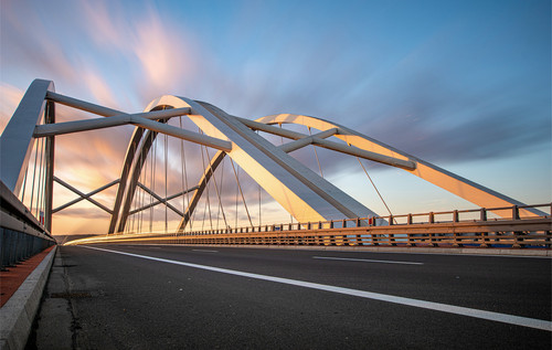Eine Stahlbrücke mit geschwungenen Trägern überspannt eine Straße, die unter der Konstruktion verläuft. Die Brückenelemente bestehen aus miteinander verbundenen Metallstreben, im Hintergrund ist ein Himmel bei Sonnenuntergang zu sehen.