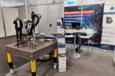 Ein Messestand der SLV Halle mit einem kollaborativen Schweißroboter auf einem Werkstücktisch. Im Hintergrund sind Rollups mit Informationen zu Aus- und Weiterbildung, Forschung und 3D-Druck-Prozessentwicklung zu sehen. Ein Mann telefoniert.