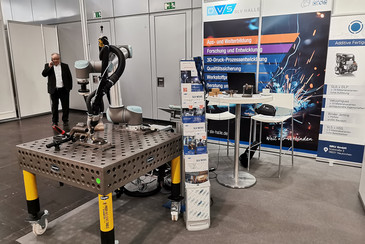 Ein Messestand der SLV Halle mit einem kollaborativen Schweißroboter auf einem Werkstücktisch. Im Hintergrund sind Rollups mit Informationen zu Aus- und Weiterbildung, Forschung und 3D-Druck-Prozessentwicklung zu sehen. Ein Mann telefoniert.