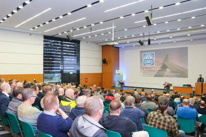 Der große Veranstaltungssaal der SLV Halle ist gefüllt mit Menschen. Vorne steht ein Mann am Pult und präsentiert eine Folie.