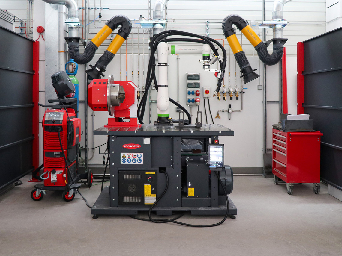 Ein Schweißarbeitsplatz mit mehreren technischen Geräten steht zwischen schwarzen Schutzwänden in einer Werkhalle. Sichtbar sind zwei Absaugarme, ein Schweißgerät, ein roter Werkzeugwagen und eine zentrale Vorrichtung mit angeschlossenen Kabeln und Schläuchen.