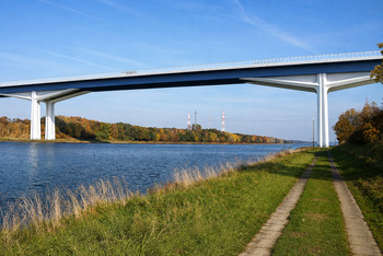 Eine hohe Straßenbrücke überquert ein breites Gewässer und wird von großen Betonpfeilern getragen. Am Ufer verläuft ein zweispuriger Fahrweg durch eine grasbewachsene Landschaft mit Bäumen, im Hintergrund sind Strommasten und rot weiße Türme zu sehen.