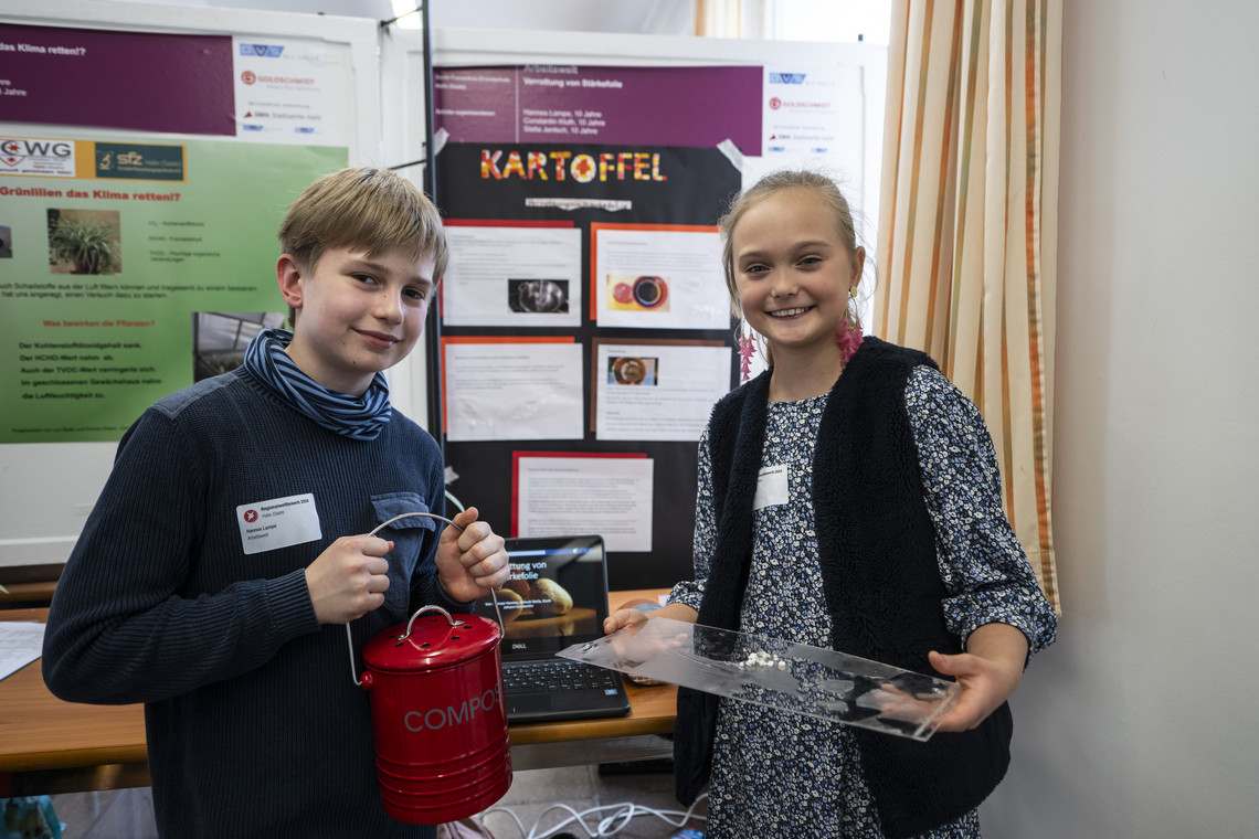 Zwei Kinder präsentieren stolz ihr Forschungsprojekt. Der Junge hält einen roten Behälter mit Kabeln, das Mädchen eine durchsichtige Platte. Im Hintergrund ein Poster mit dem Titel „Kartoffel“.