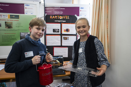 Zwei Kinder präsentieren stolz ihr Forschungsprojekt. Der Junge hält einen roten Behälter mit Kabeln, das Mädchen eine durchsichtige Platte. Im Hintergrund ein Poster mit dem Titel „Kartoffel“.