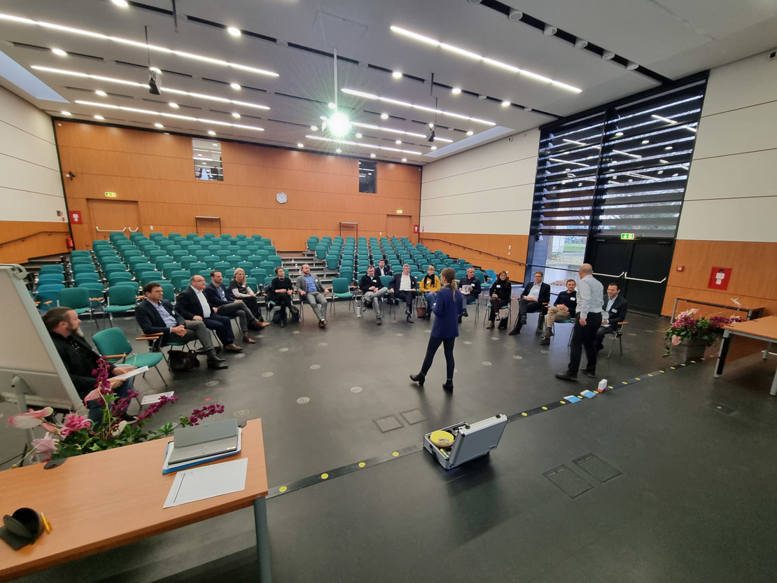 Personen sitzen in einem großen Hörsaal in offener Kreisform, während zwei Vortragende im vorderen Bereich stehen; leere Sitzreihen im Hintergrund, Blumen und Technik auf dem Podium im Vordergrund.