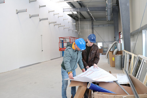 Zwei Männer mit blauen Schutzhelmen betrachten Baupläne in einer Industriehalle im Ausbauzustand, umgeben von Baumaterial, im Hintergrund Hebebühne und Lüftungsrohre