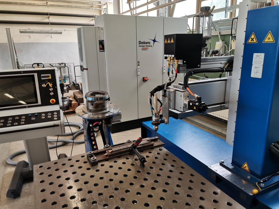 Automatisierte Laserauftragsschweißanlage mit großem Schweißtisch, Bedienterminal und zentralem Gehäuse der Firma Deloro; verschiedene Werkzeuge und Bauteile sind auf dem Tisch positioniert.