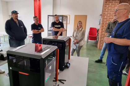 Vier Männer und eine Frau stehen in einer Werkhalle an einem technischen Arbeitsplatz mit mehreren 3D-Druckern auf einem Arbeitstisch. Eein weiterer Mann steht rechts daneben und erklärt etwas, während die anderen zuhören. Im Hintergrund sind Stühle zu sehen.