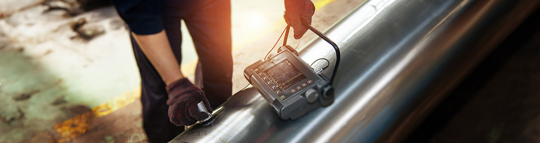 Prüfperson führt eine Ultraschallprüfung an einer Metallwelle durch, während die Messergebnisse auf einem tragbaren Gerät angezeigt werden.