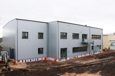 Neubau eines zweigeschossigen Gebäudes mit grauer Metallfassade und Außentreppe, Erdarbeiten rund um das Gebäude noch im Gange, Minibagger links im Bild