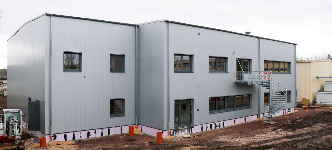 Neubau eines zweigeschossigen Gebäudes mit grauer Metallfassade und Außentreppe, Erdarbeiten rund um das Gebäude noch im Gange, Minibagger links im Bild