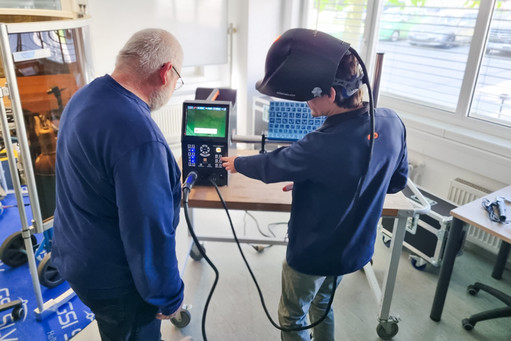 Zwei Männer stehen an einem Elektronikarbeitsplatz, eine Person bedient mit beiden Händen Werkzeuge vor einem Monitor, während die andere daneben zuschaut. Auf dem Tisch befinden sich Messgeräte, Kabel und Bauteile, im Hintergrund ist eine Werkstattumgebung mit Fenstern und weiteren Geräten zu sehen.