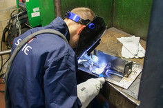 Eine Person in blauer Schutzkleidung mit Schweißhelm und Handschuhen sitzt an einem Schweißtisch und arbeitet konzentriert an einem Werkstück; im Hintergrund eine grüne Schutzwand und ein Schweißgerät.