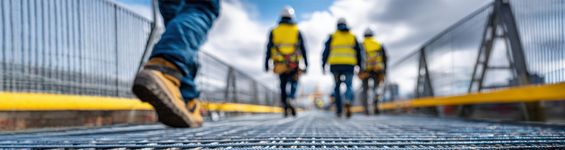 Vier Personen bewegen sich über einen Metallgitterrost auf einer Baustelle, im Vordergrund ist ein Schuh in Nahaufnahme zu sehen, im Hintergrund gehen drei Personen mit Warnwesten und Schutzhelmen von der Kamera weg. Seitlich verlaufen Geländer, die Oberfläche besteht aus einem engmaschigen Stahlgitter, das als Laufweg dient.