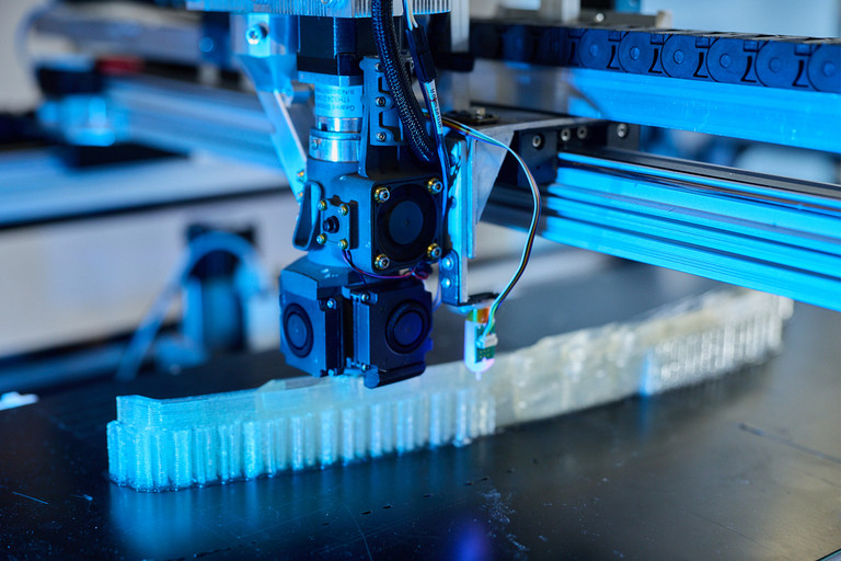 Nahaufnahme eines 3D-Druckkopfs, der ein längliches, helles Kunststoffbauteil schichtweise auf einer schwarzen Druckplattform fertigt. Im Hintergrund sind Führungsschienen und Kabel der Druckanlage zu sehen.