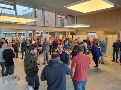 Großes Foyer mit vielen Besuchenden beim informellen Austausch während einer Veranstaltungspause; Stehtische mit Getränken und Tellern im Vordergrund. Ca. 50 Personen, überwiegend Männer.