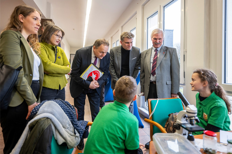 Mehrere Erwachsene stehen um einen Tisch, an dem zwei Jugendliche sitzen und technische Unterlagen sowie Bauteile betrachten, während eine Person im Hintergrund fotografiert und der Austausch in einem hellen Raum mit großen Fenstern stattfindet.