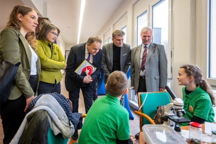 Mehrere Erwachsene stehen um einen Tisch, an dem zwei Jugendliche sitzen und technische Unterlagen sowie Bauteile betrachten, während eine Person im Hintergrund fotografiert und der Austausch in einem hellen Raum mit großen Fenstern stattfindet.