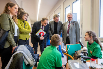 Mehrere Erwachsene stehen um einen Tisch, an dem zwei Jugendliche sitzen und technische Unterlagen sowie Bauteile betrachten, während eine Person im Hintergrund fotografiert und der Austausch in einem hellen Raum mit großen Fenstern stattfindet.