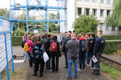 Gruppe von Personen steht im Außenbereich eines Geländes und hört einer erklärenden Person zu, im Hintergrund industrielle Anlagen und Gebäude