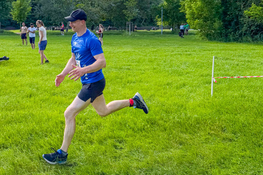 Ein Mann in blauem Laufshirt und kurzer Sporthose läuft über eine Wiese entlang einer markierten Strecke. Im Hintergrund stehen mehrere Personen auf der Wiese, daneben sind Fahrräder und Bäume zu sehen.