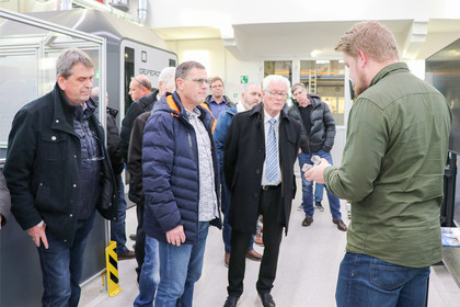 Mehrere Männer in Winterjacken stehen in einer Werkhalle um einen Mann, der ein Metallbauteil erklärt, im Hintergrund industrielle Anlagen und Maschinen.