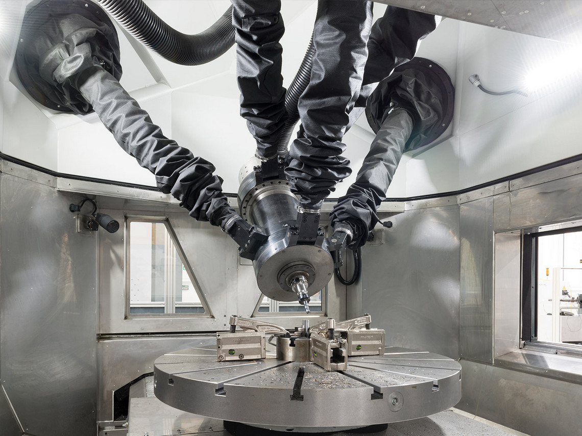 Eine Spindel mit 5 Bewegungsachsen und 2 Zusatzachsen mit Faltenbalgabdeckung in einer geschlossenen METROM-Zelle richtet sich auf ein eingespanntes Metallwerkstück auf einem Rundtisch.